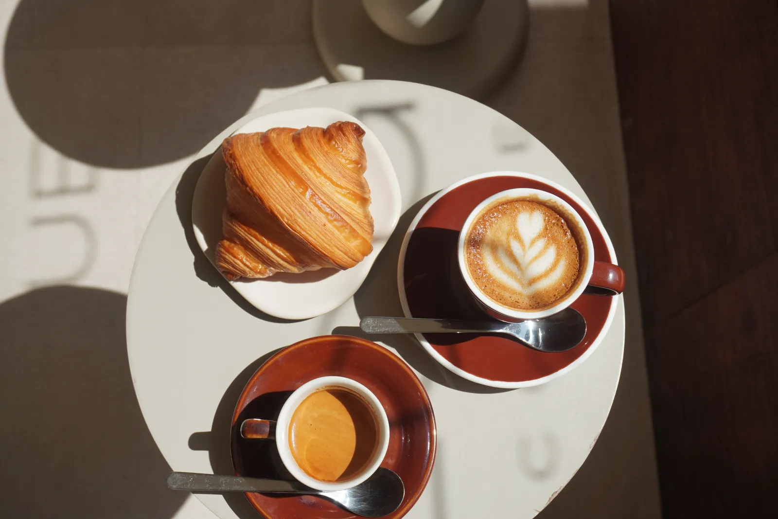 Overhead: a croissant, a cappuccino and an espresso on a white café table in raking morning light.