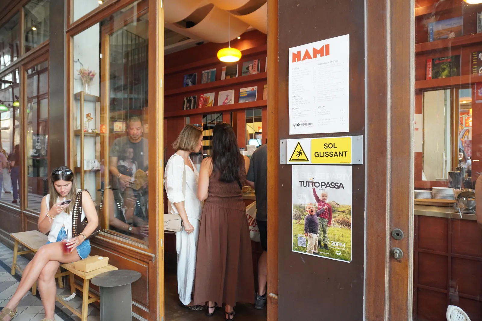 The open doorway of Nami: guests at the counter inside, a woman on the bench out front, the NAMI menu posted by the door.